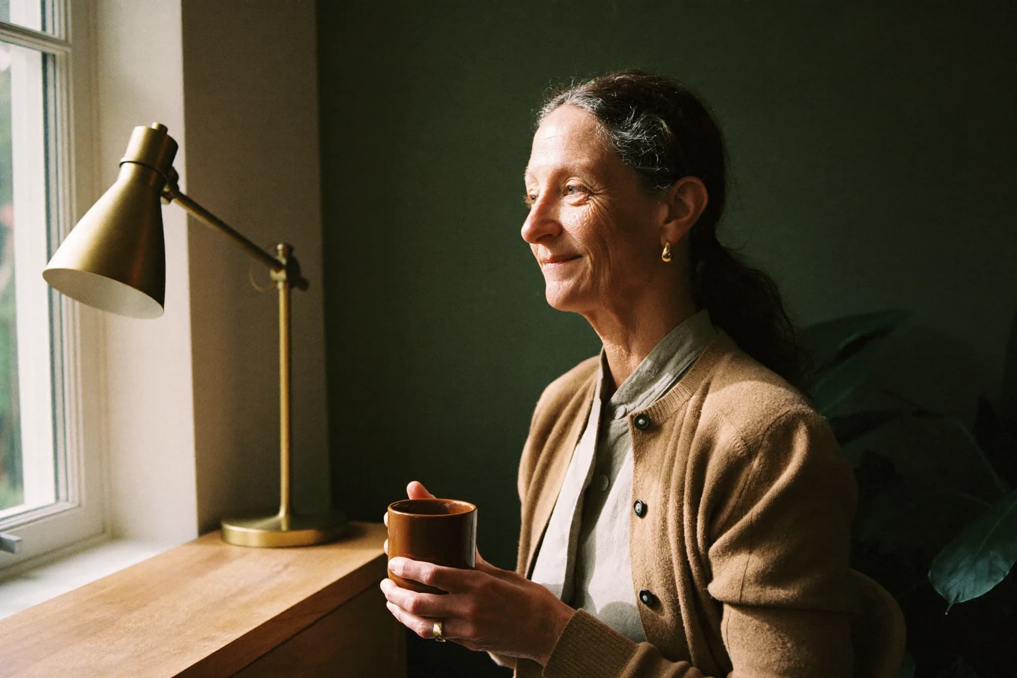 Een vrouw met een mok koffie bij het raam, in warm ochtendlicht.